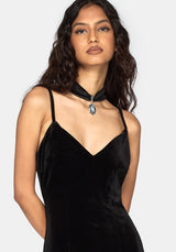 A woman modeling a black velvet slip dress with thin straps and a matching black choker necklace featuring a silver-toned pendant, set against a plain light gray background.