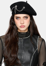A woman wearing a black beret with silver chain and ring embellishments, paired with a black mesh and faux-leather top, against a light gray studio background.
