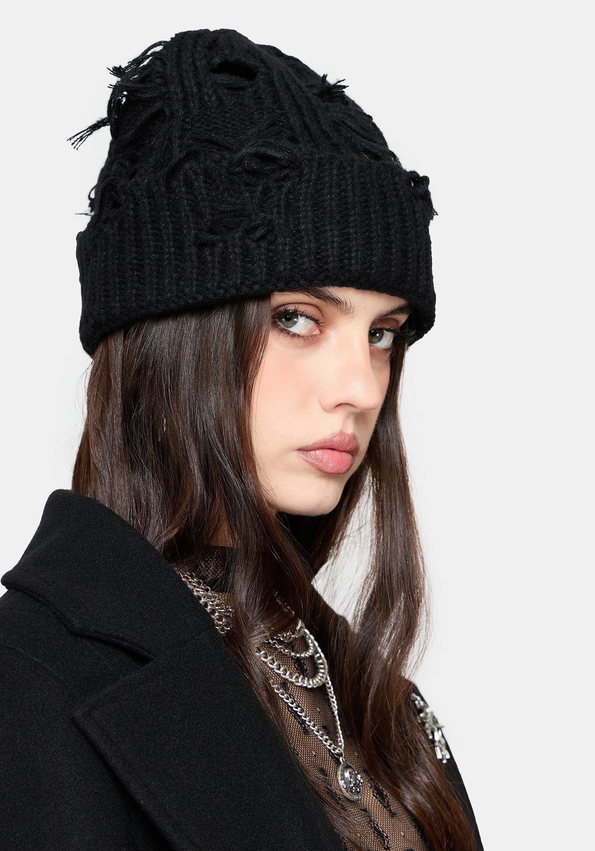 A woman wearing a black cable-knit beanie with distressed frayed details and a black coat, shown in a close-up portrait against a light gray background.