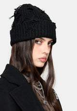 A woman wearing a black cable-knit beanie with distressed frayed details and a black coat, shown in a close-up portrait against a light gray background.