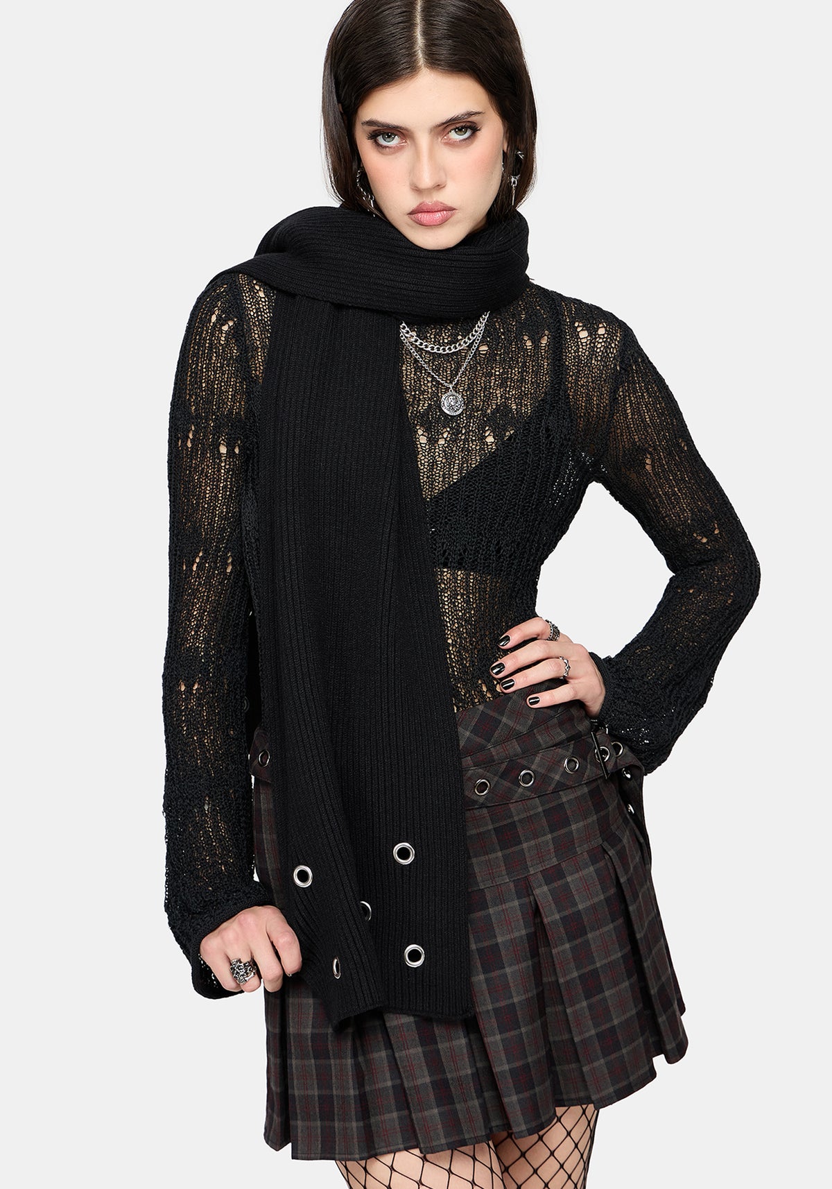 A model wearing a black knit wrap scarf, a sheer black lace long-sleeve top, and a dark green and black plaid pleated mini skirt with silver grommets, set against a plain light gray background.