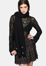 A model wearing a black knit wrap scarf, a sheer black lace long-sleeve top, and a dark green and black plaid pleated mini skirt with silver grommets, set against a plain light gray background.