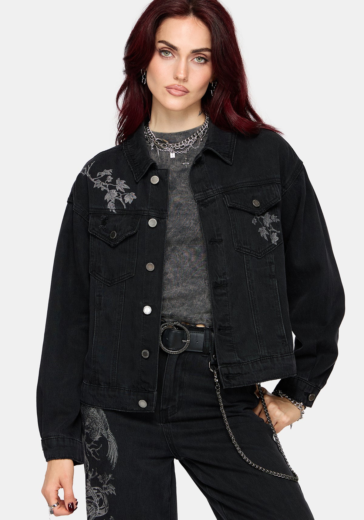 A model with dark red hair wearing a black denim jacket with grey floral embroidery on the chest, layered over a gray textured top and black jeans.