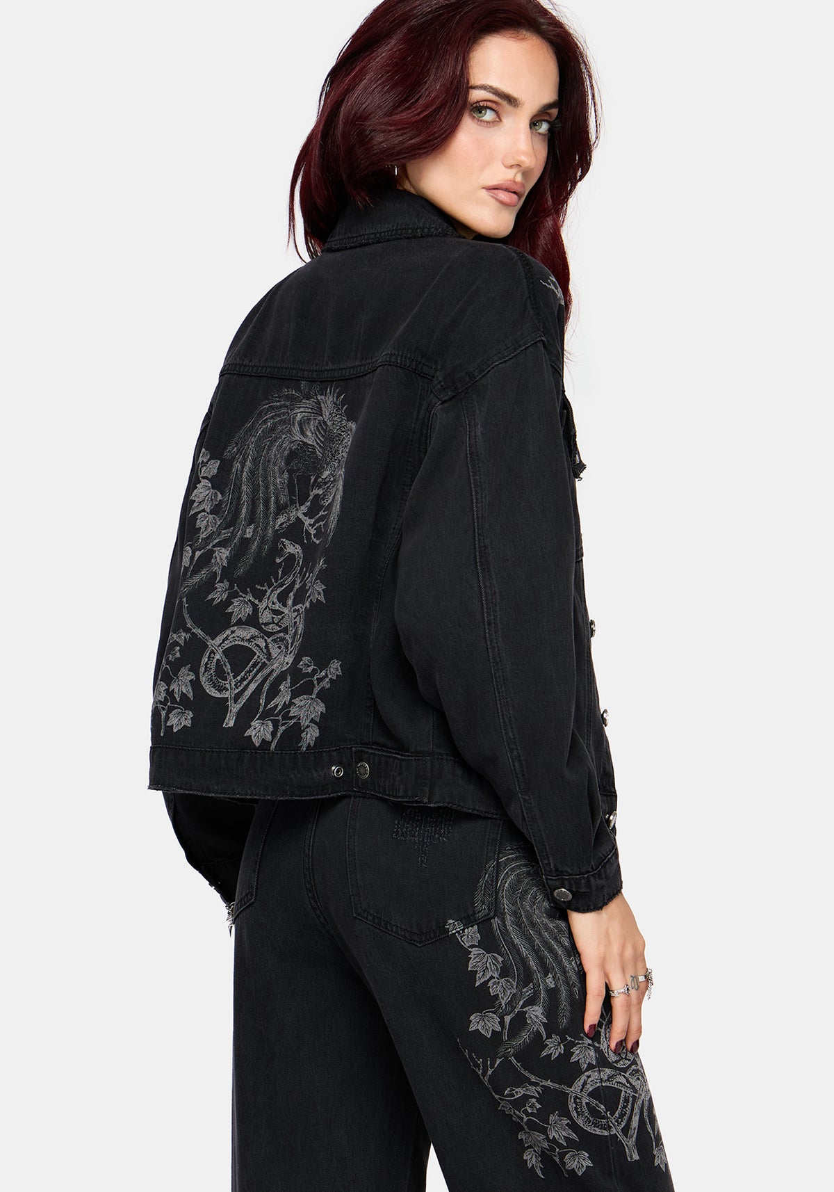 A woman wearing a black denim jacket with faded grey graphic prints on the back and upper thigh, paired with black jeans, photographed against a light gray studio background.