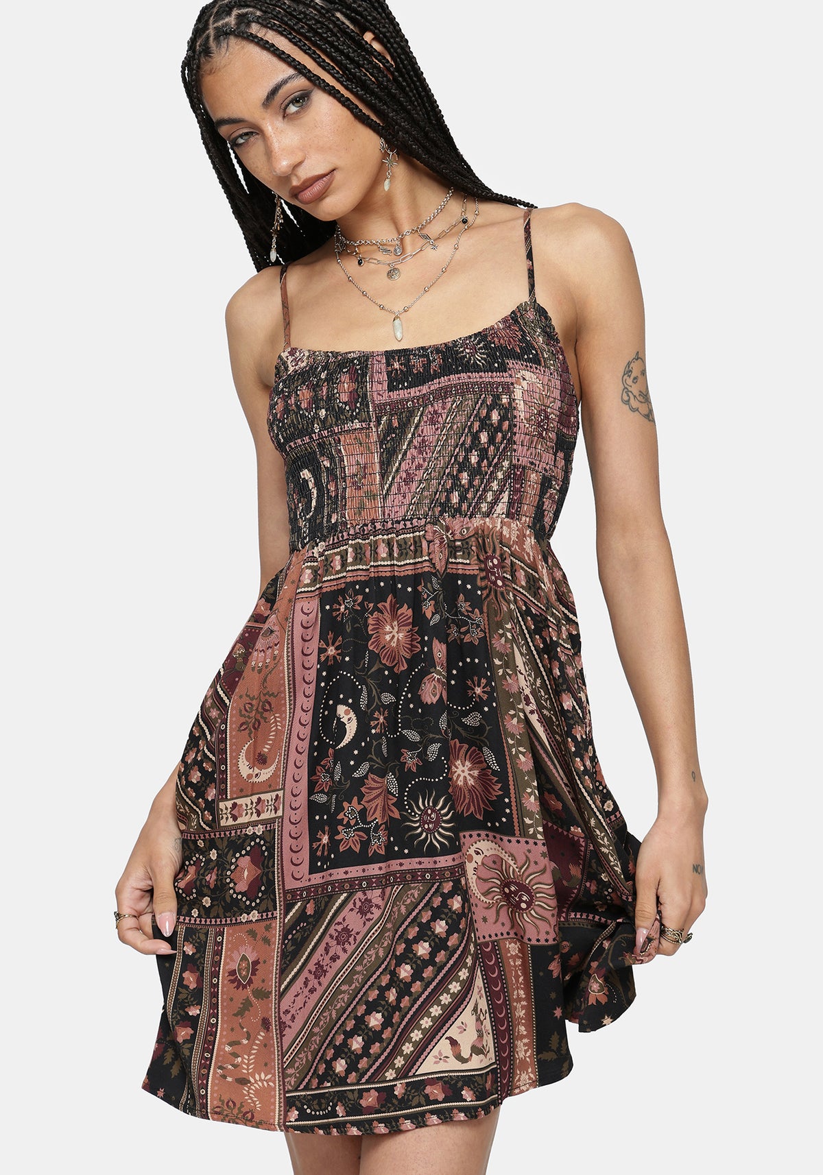 A woman modeling a sleeveless mini dress with thin straps and an allover patchwork-style bohemian print in dusty pink, rust, beige, and black, against a light gray studio background.