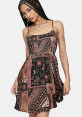A woman modeling a sleeveless mini dress with thin straps and an allover patchwork-style bohemian print in dusty pink, rust, beige, and black, against a light gray studio background.