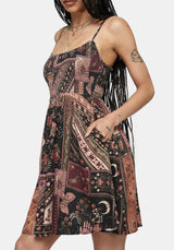 A woman wearing a sleeveless spaghetti-strap mini dress with a bohemian patchwork print in dusty pink, rust, brown, and black, shown against a light gray studio background.