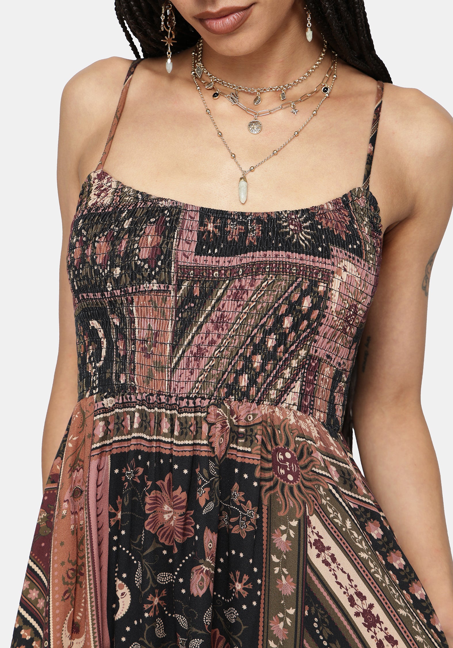 A close-up of a woman wearing a sleeveless patchwork-print dress with thin straps and an elasticated waist, shown against a plain light background. The dress has a multicolor print with pink, black, and brown earthy tones.