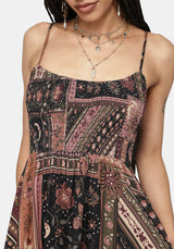 A close-up of a woman wearing a sleeveless patchwork-print dress with thin straps and an elasticated waist, shown against a plain light background. The dress has a multicolor print with pink, black, and brown earthy tones.