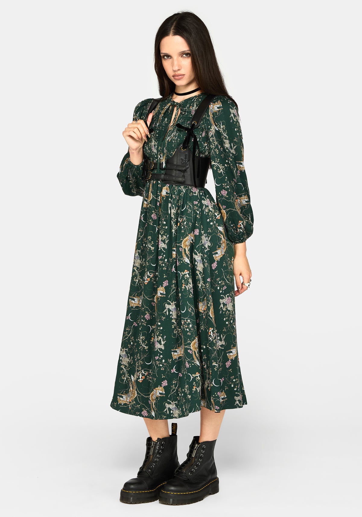 A model wearing a dark green midi dress with a woodland animal print, styled with a black faux-leather corset-style waist belt and black combat boots, against a light gray studio background.