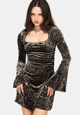 A woman modeling a black crushed velvet long-sleeve dress with a square neckline, ruched bodice, and flared bell sleeves against a light gray background.