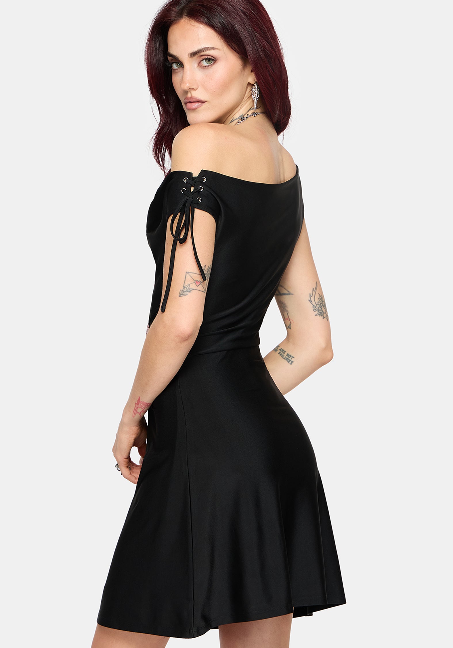 A woman modeling a black off-the-shoulder skater dress with lace-up detailing on the back, shown against a light gray studio background.