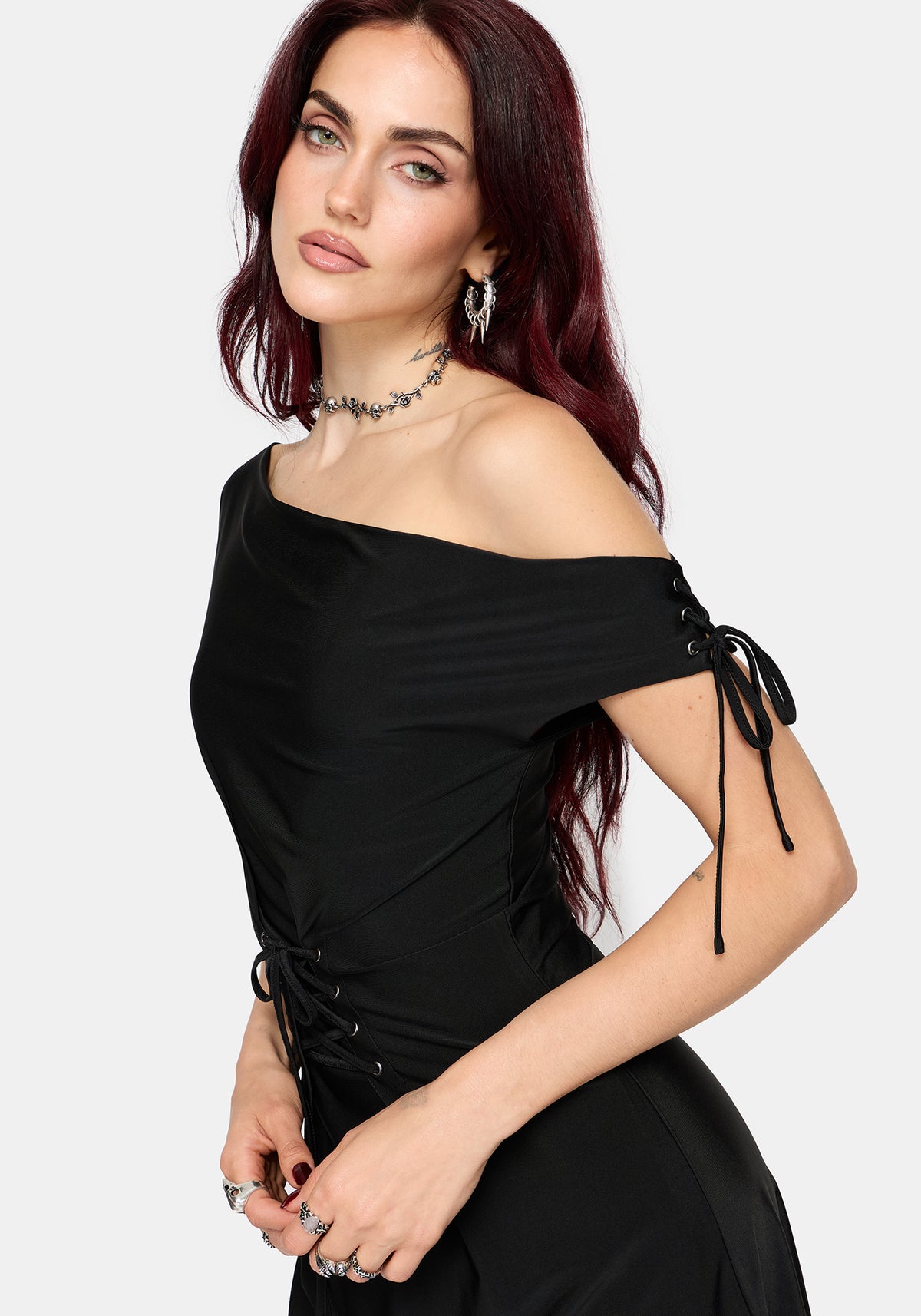 A woman modeling a black off-the-shoulder dress with side lace-up details, photographed against a light gray studio background.