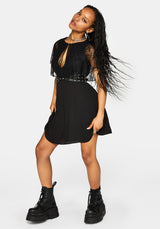 A woman modeling a black mini dress with sheer flutter sleeves, a plunging keyhole neckline, and a studded waist belt, paired with chunky black combat boots against a light gray studio background.