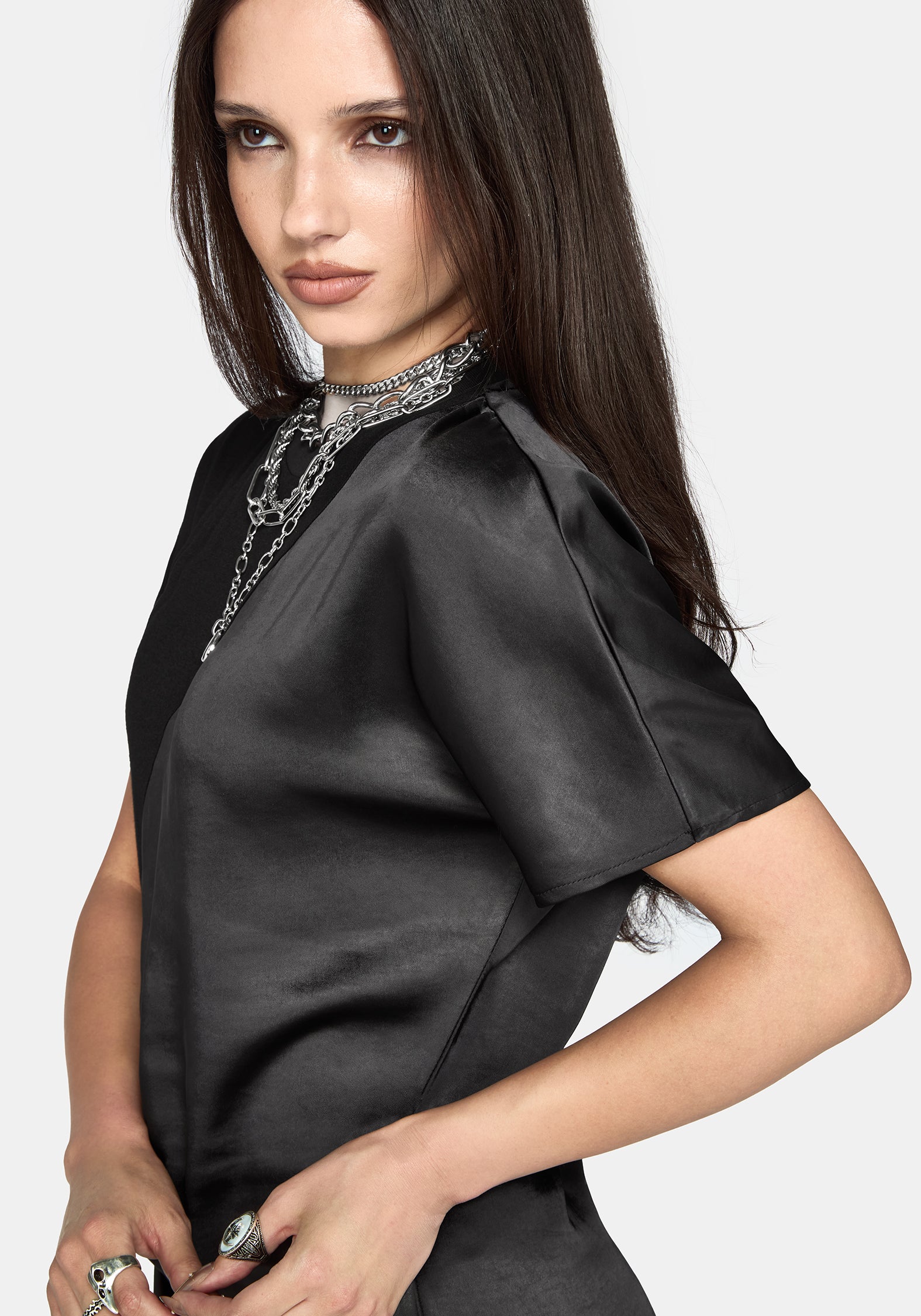 A woman wearing a black short-sleeve satin dress with a high mock neck and silver chain detailing, photographed against a light gray studio background.
