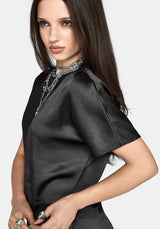 A woman wearing a black short-sleeve satin dress with a high mock neck and silver chain detailing, photographed against a light gray studio background.
