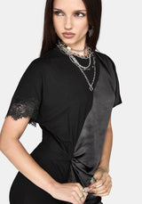 A woman wearing a black short-sleeve dress with a satin diagonal panel, lace trim on the sleeve, and a twisted knot at the waist, styled with layered silver chain necklaces against a light gray background.