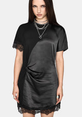 A woman modeling a black satin-like short-sleeve dress with lace trim at the sleeve cuffs and hem, layered with silver chain necklaces, against a light gray studio background.
