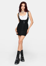 A model wearing a black sleeveless mini dress with a white ribbed tank top underneath, accessorized with layered necklaces and black lace-up platform boots, set against a plain light gray background.
