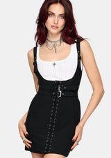 A woman modeling a black pinafore-style mini dress with a white ribbed tank top underneath. The dress features shoulder straps, a self-tie waist belt with silver buckles, and lace-up detailing down the front.