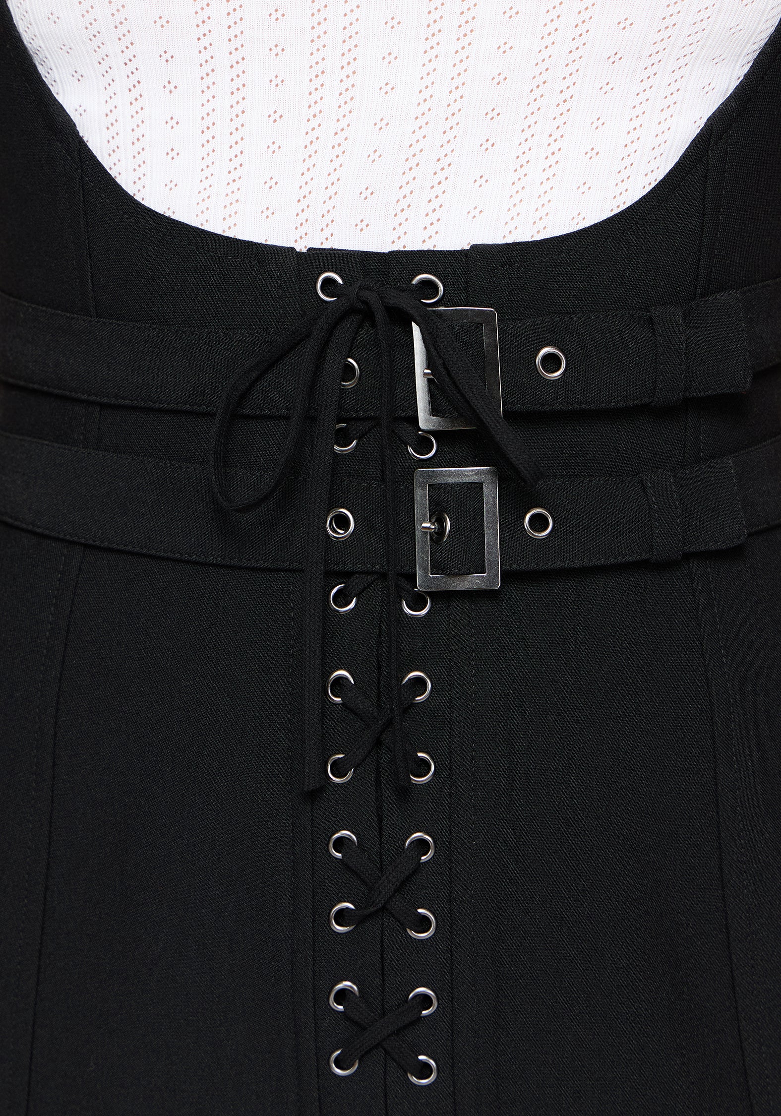 Close-up of a black dress featuring a lace-up front with silver metal eyelets and two horizontal black straps with square silver buckles, set against a white textured knit top.