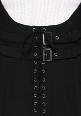 Close-up of a black dress featuring a lace-up front with silver metal eyelets and two horizontal black straps with square silver buckles, set against a white textured knit top.