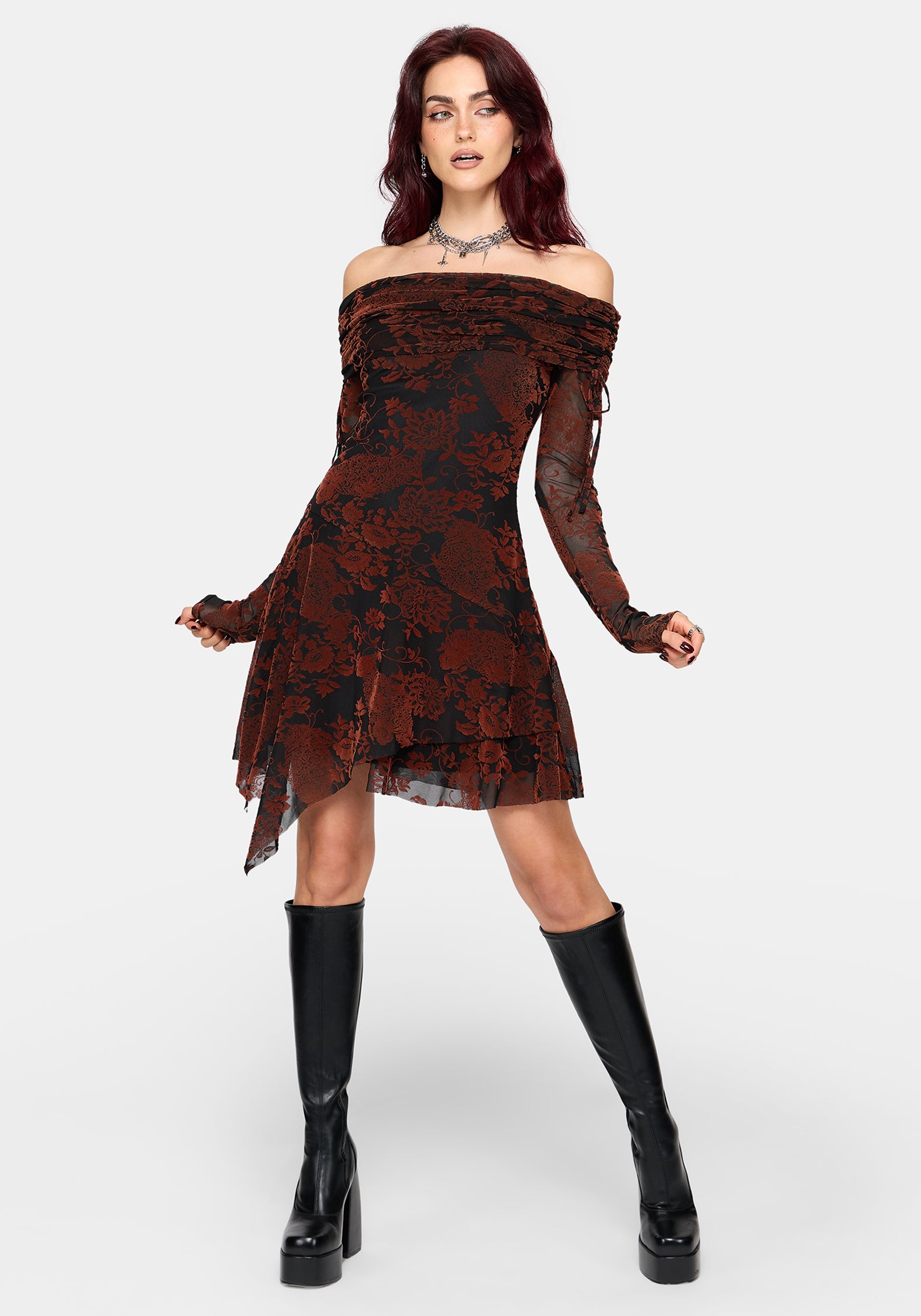 A model wearing a burnt orange off-the-shoulder floral mesh dress with sheer long sleeves, paired with black knee-high platform boots, against a light gray studio background.