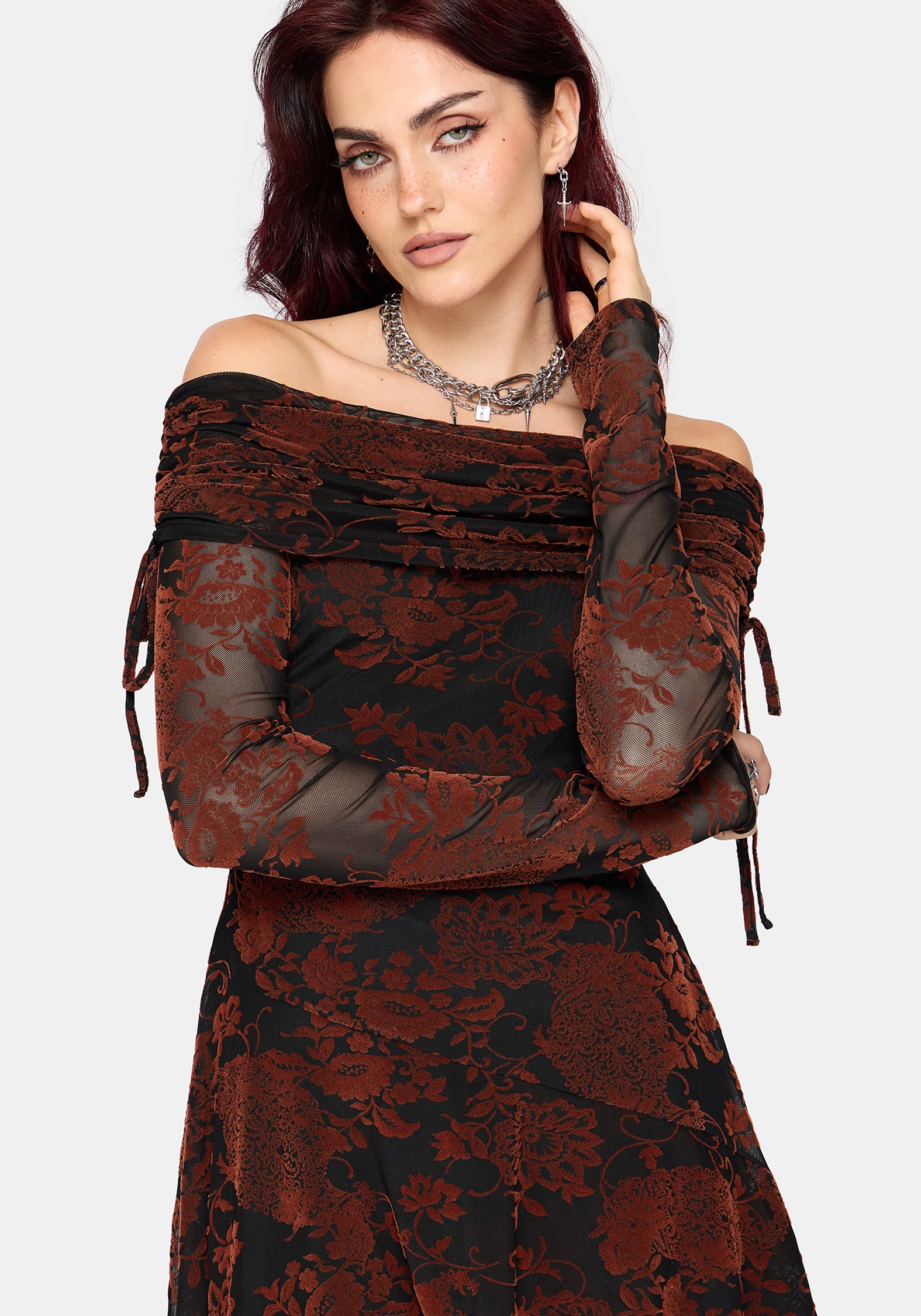 A woman modeling an off-the-shoulder long-sleeve dress with a black and rust-brown floral jacquard pattern, sheer mesh sleeves, and side tie details, set against a plain light gray background.