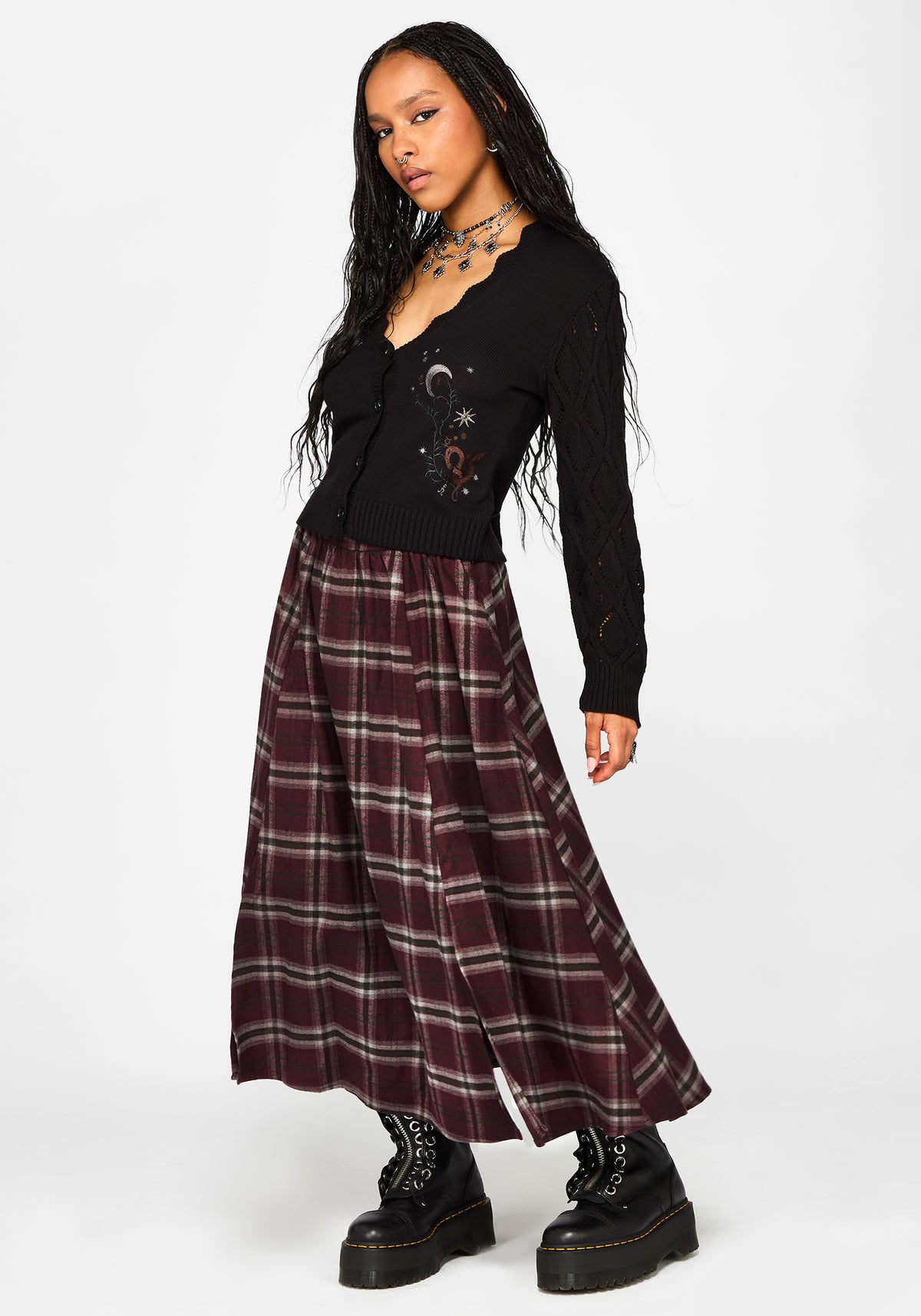 A model wearing a black long-sleeve jumper with celestial embroidery and a long burgundy plaid skirt, paired with black combat boots, against a plain white studio background.