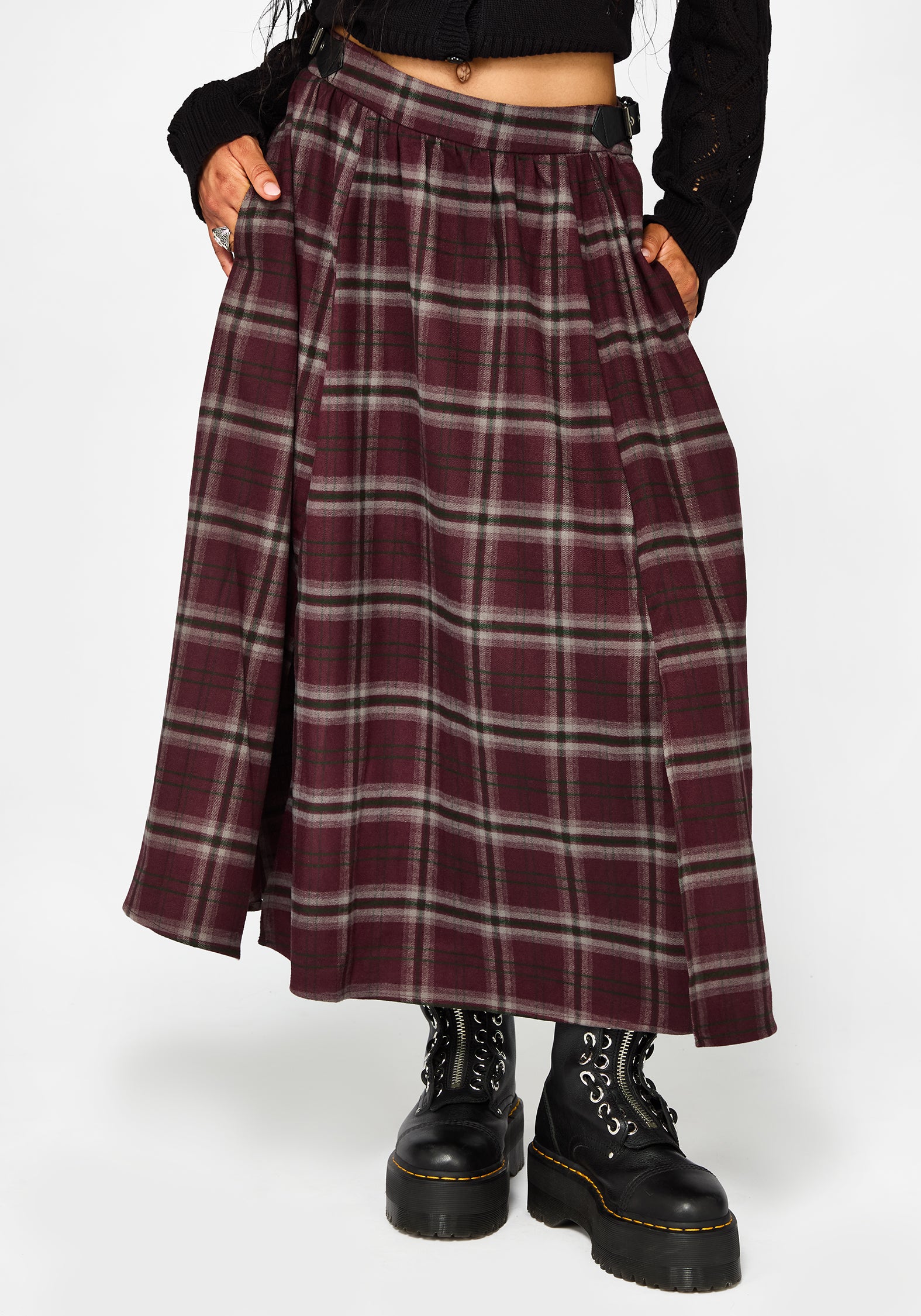 A person wearing a pleated A-line skirt in a burgundy and gray plaid pattern, styled with a black crop top and black combat boots against a light gray background.