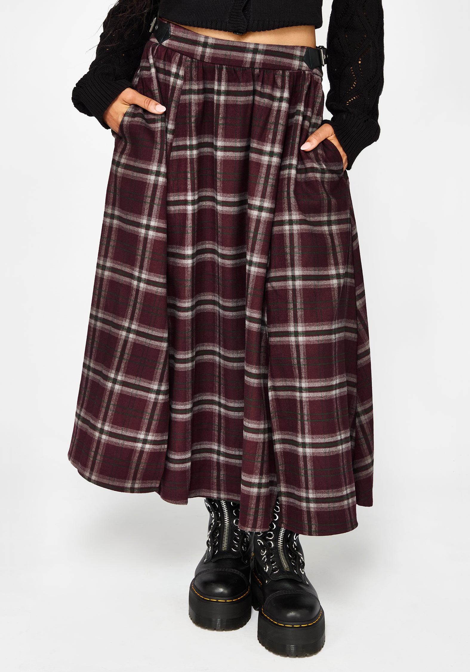 A person wearing a long pleated plaid skirt in burgundy, black, and white, paired with black platform boots and a black cropped top, shown against a light gray studio background.