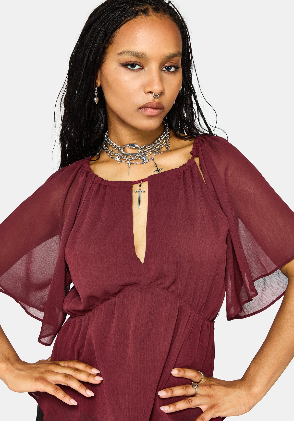 A woman wearing a sheer, flowy burgundy top with flutter sleeves and a split neckline with a keyhole cutout, accessorized with layered silver necklaces, against a light gray background.