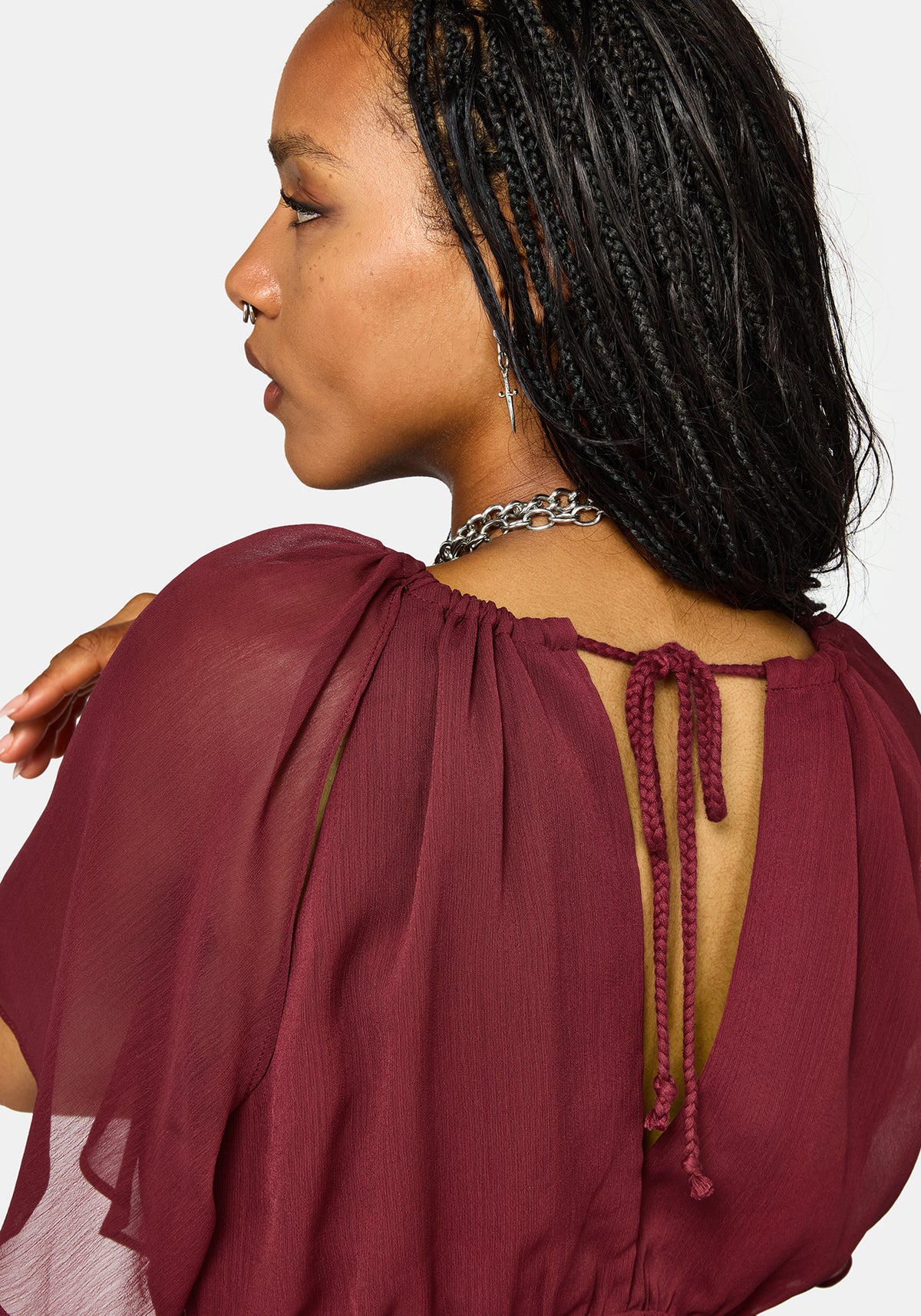 A close-up side and back view of a woman wearing a sheer burgundy top with a tie-back open back and braided hair against a light gray background.