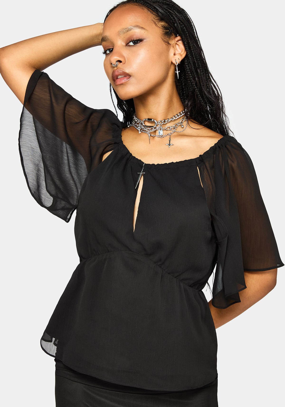 A woman wearing a black sheer flutter-sleeve peplum top with a keyhole cutout at the neckline, styled with layered silver necklaces against a light gray background.