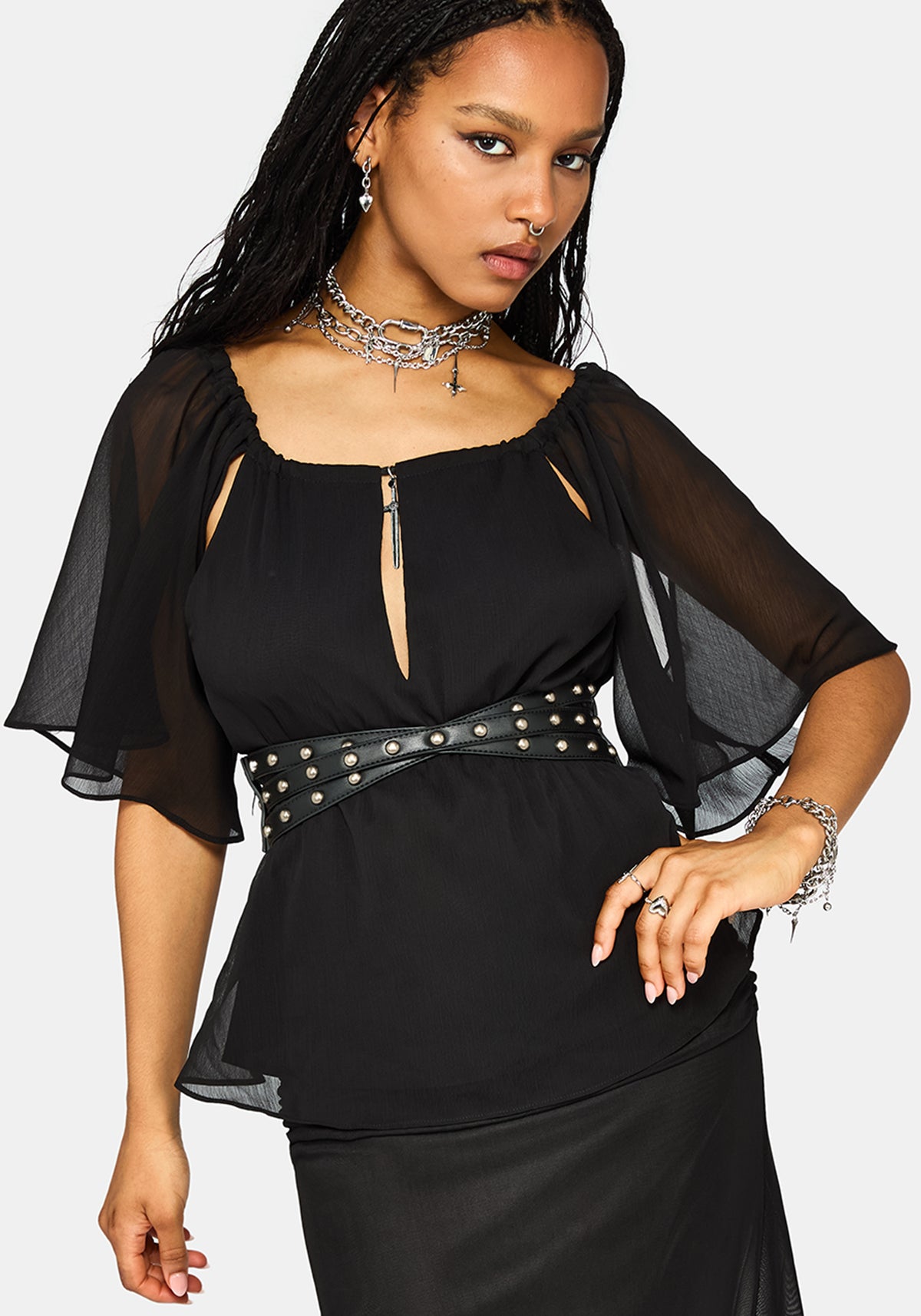 A woman modeling a black blouse with sheer flutter sleeves, a scoop neckline with a front zipper cutout, a wide studded black belt, and a matching black skirt, against a light gray background.