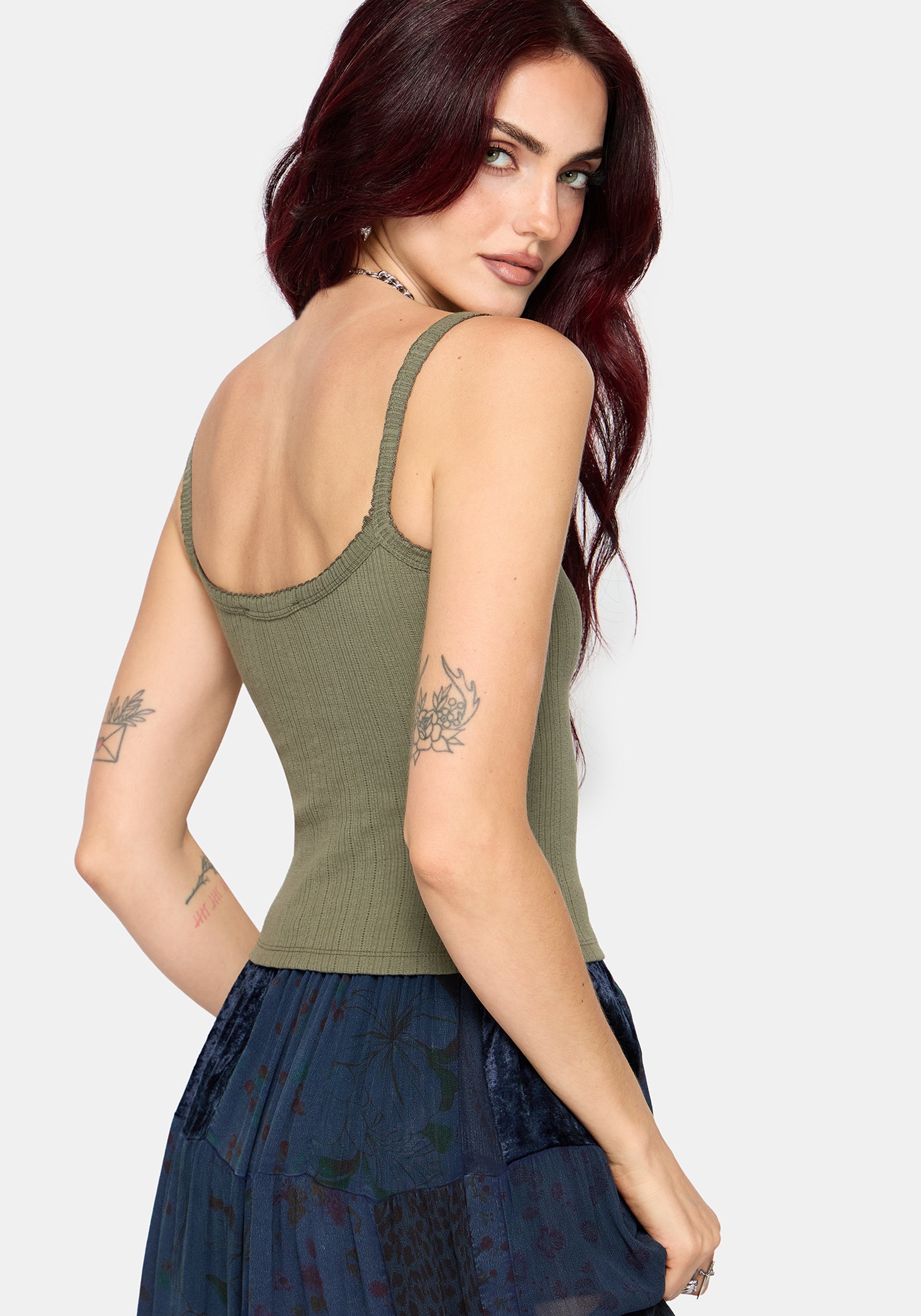 Woman wearing a khaki sleeveless vest with thin straps, cropped length and blue butterfly embroidery in center against a white studio background