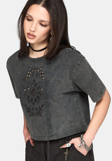 A woman wearing a dark gray washed short-sleeve crop top with a decorative embroidered panel on the front, styled with black pants and accessories against a light gray background.