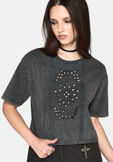 A woman wearing a dark gray washed short-sleeve crop top with a vertical panel of stud cut outs and embroidery, paired with black pants and a black choker against a light gray background.