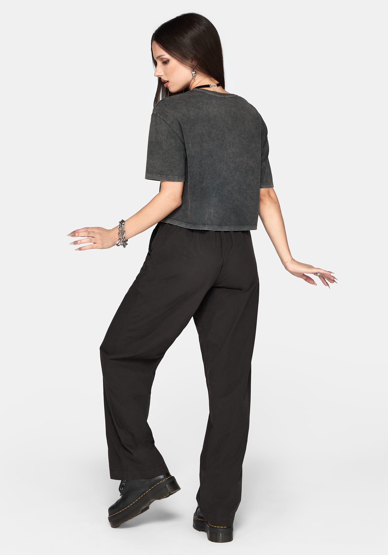 A woman posing from the back wearing a dark gray cropped t-shirt and black wide-leg trousers against a light gray background.