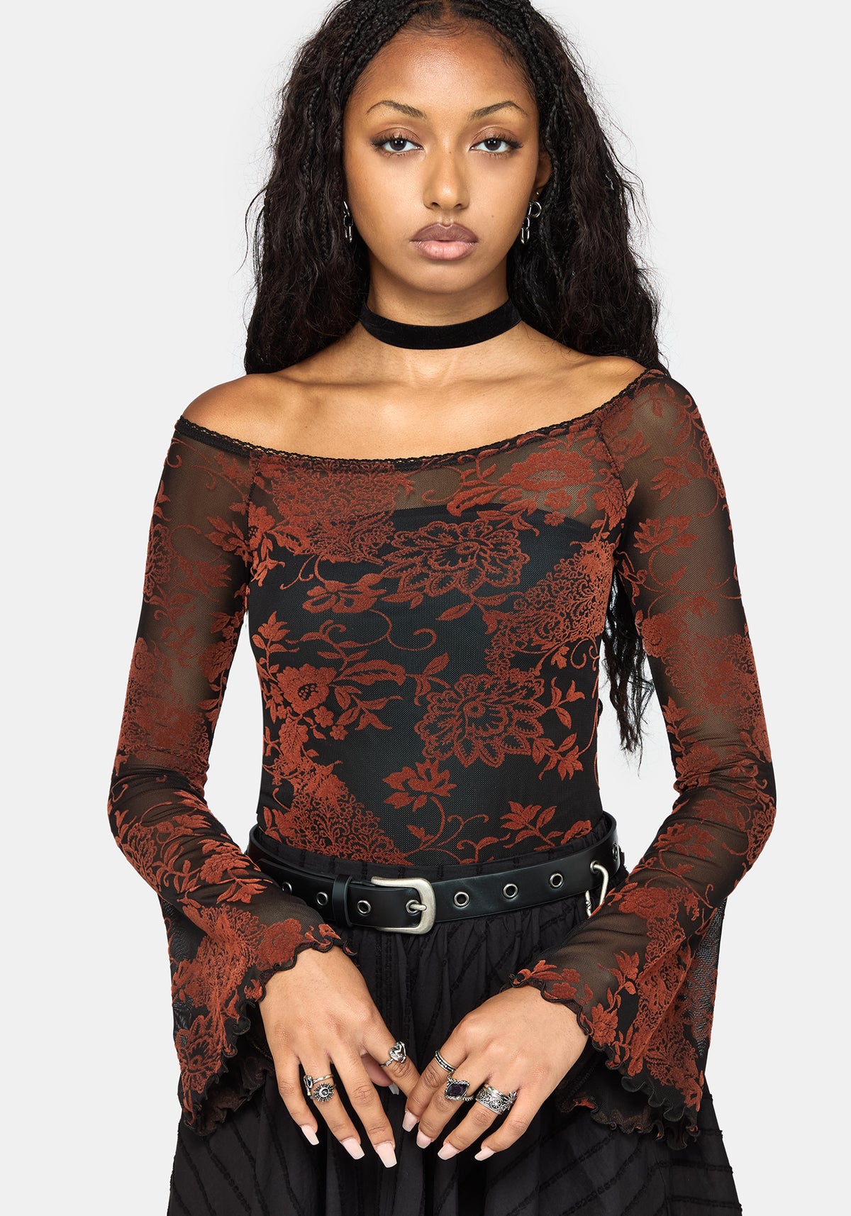 A woman modeling a black and rust red floral lace off-the-shoulder long-sleeve top with a black choker and belt, against a light gray background.